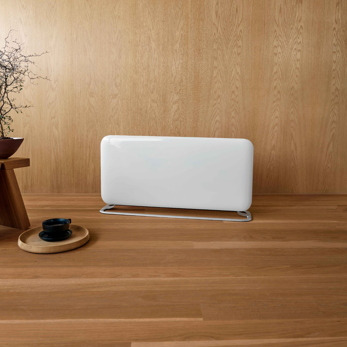 Un termosifone bianco su pavimento in legno davanti a una parete rivestita in legno. A sinistra un tavolino con una tazza di caffè.