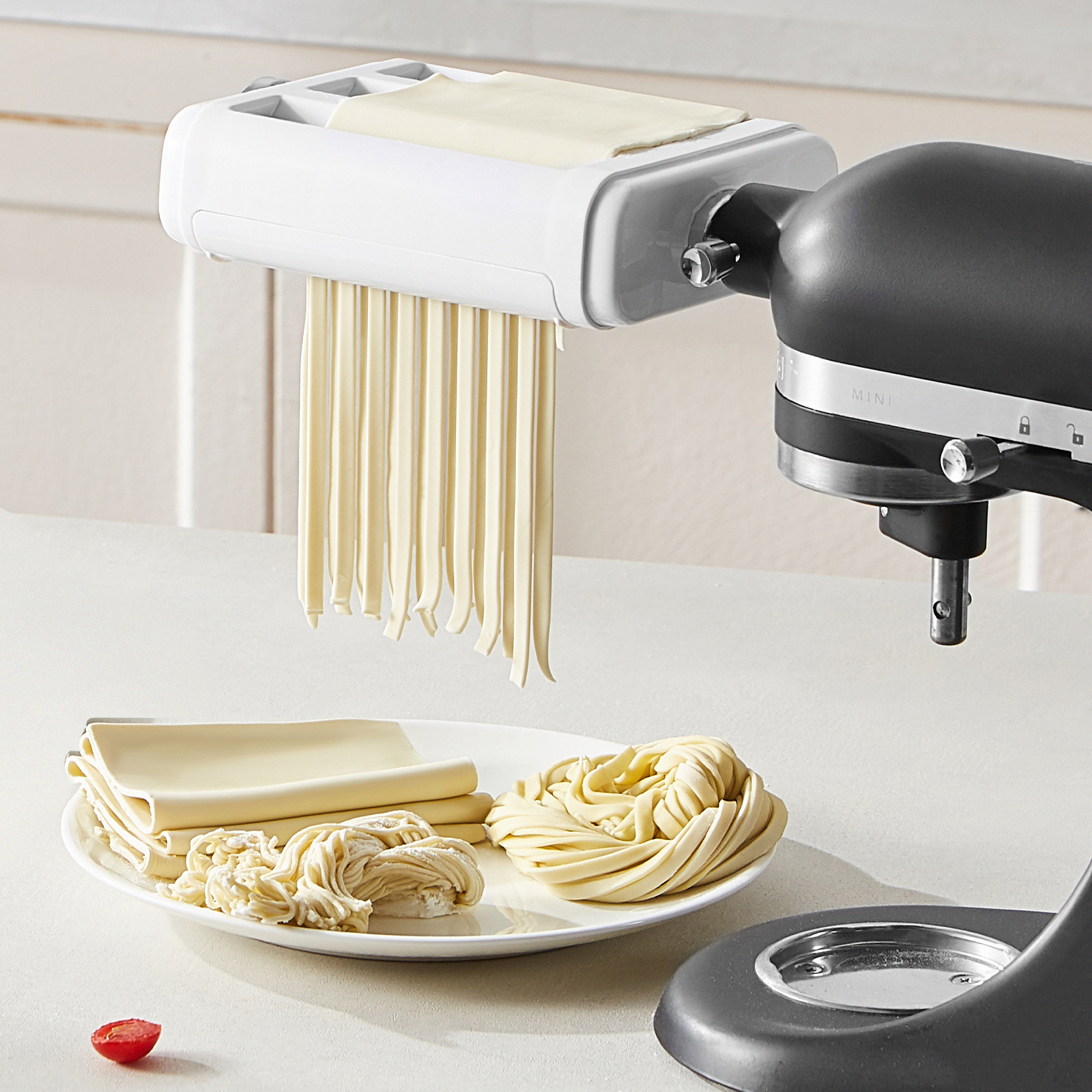 Fideos frescos cuelgan de un accesorio en una batidora negra, con láminas y nidos de pasta en un plato.