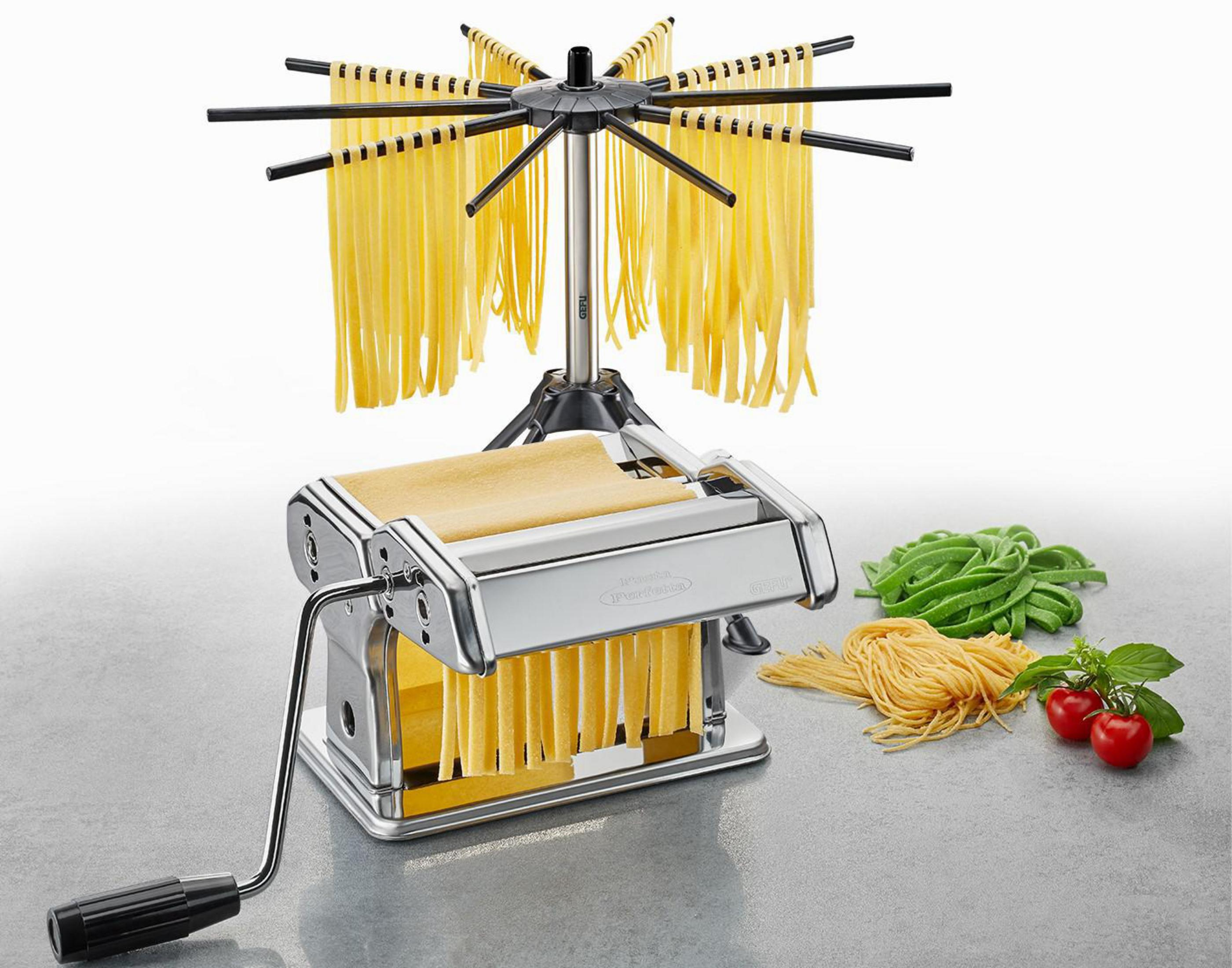 Macchina per pasta con manovella e stendino con pasta fresca e pomodori su tavolo grigio.