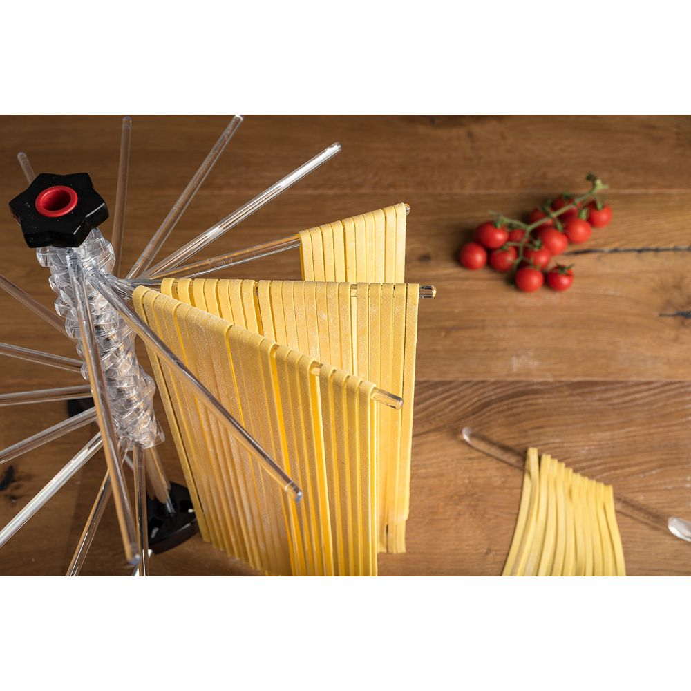 Pasta gialla fresca appesa ad asciugare su un supporto trasparente su legno, con pomodorini vicini.