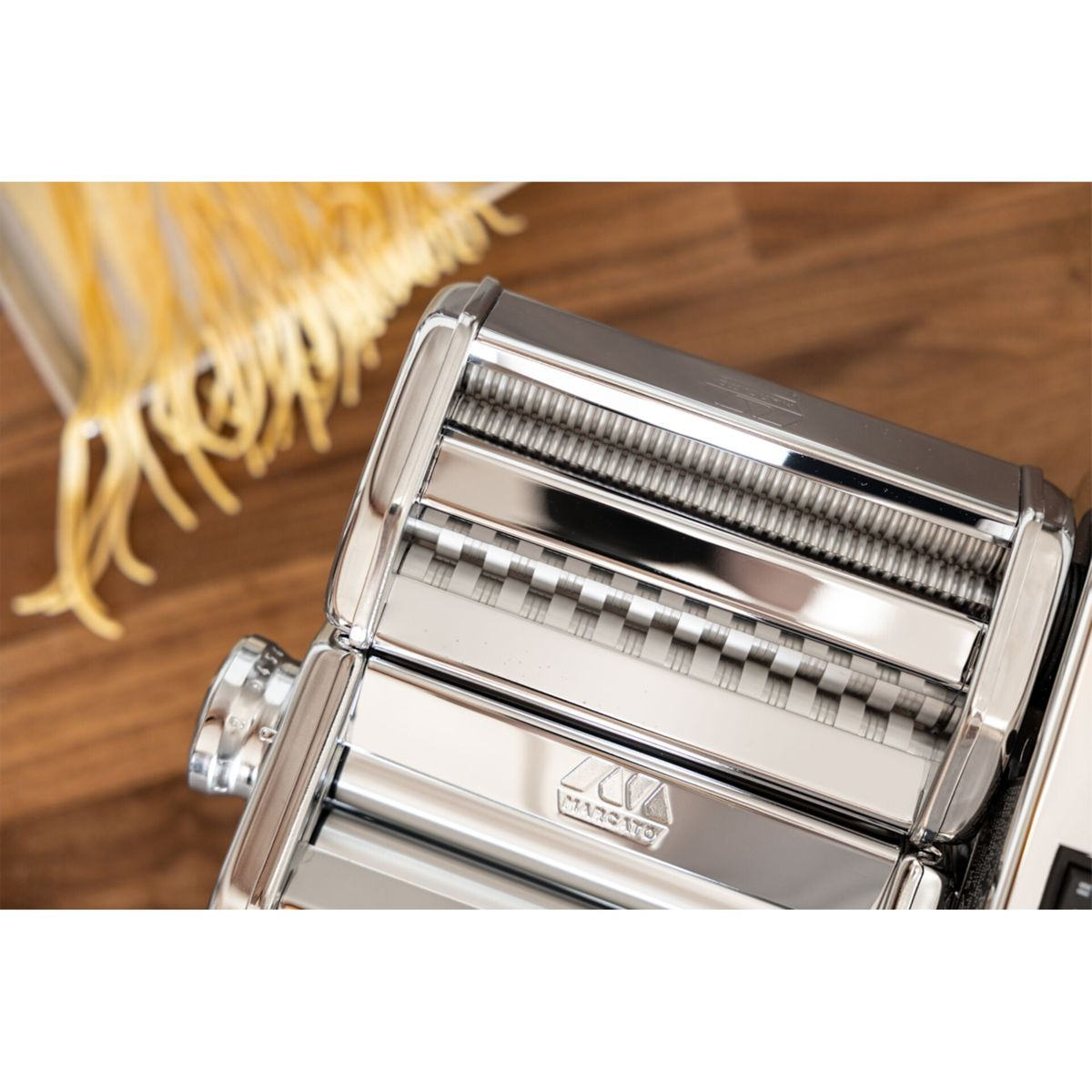 Macchina per pasta in cromo lucido su tavolo di legno con pasta fresca sullo sfondo.
