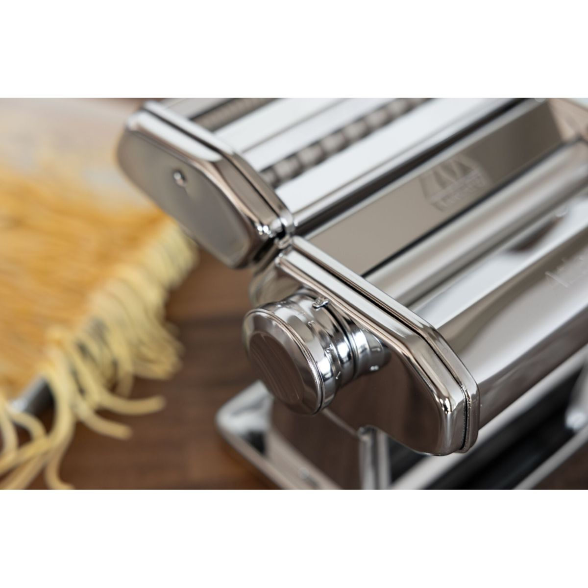 Una lucida macchina per pasta argentata si trova su una superficie di legno, con pasta fresca sullo sfondo.