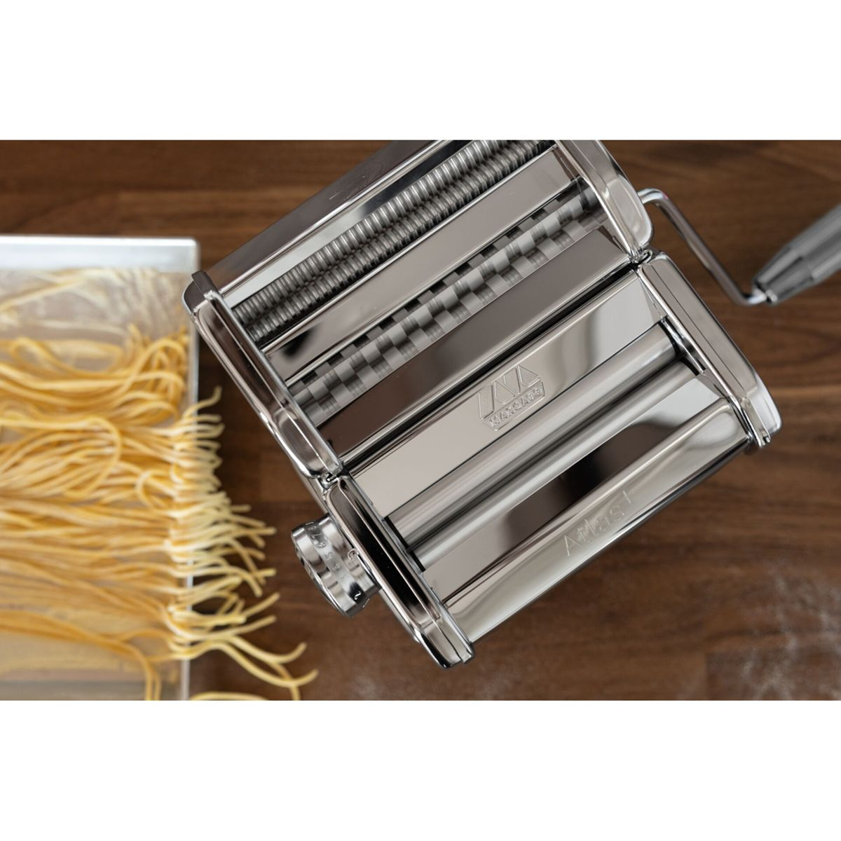 Una macchina per pasta cromata si trova su un tavolo di legno accanto a spaghetti freschi.