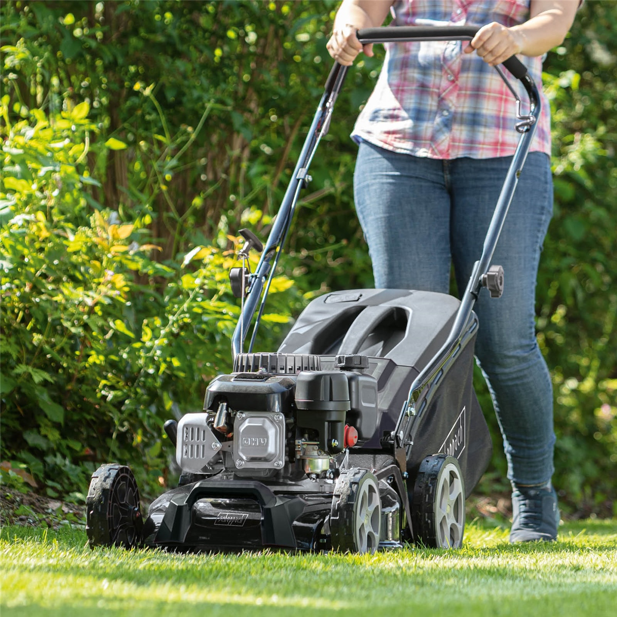 Eine Frau in Jeans und kariertem Hemd mäht mit einem schwarzen Rasenmäher den grünen Garten.