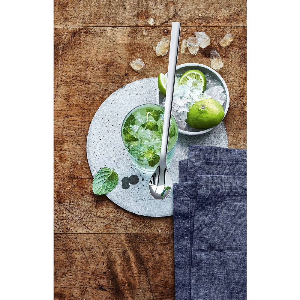 Ein langer Löffel liegt auf einem Holztisch neben einem Mojito-Glas, Limetten und blauen Servietten.