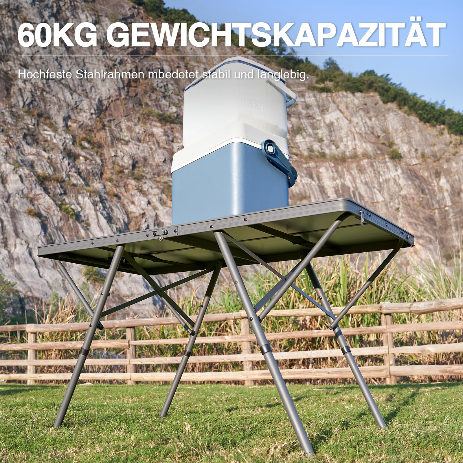 Ein grauer Campingtisch mit einer blauen Kühlbox darauf steht auf einer Wiese vor einem felsigen Hang.