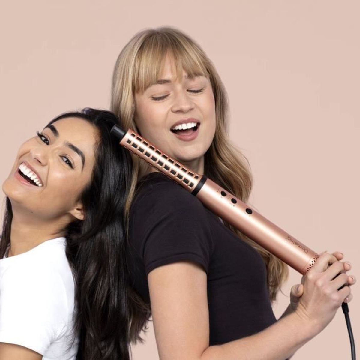 Due donne sorridenti tengono un ferro arricciacapelli oro rosa su sfondo beige.