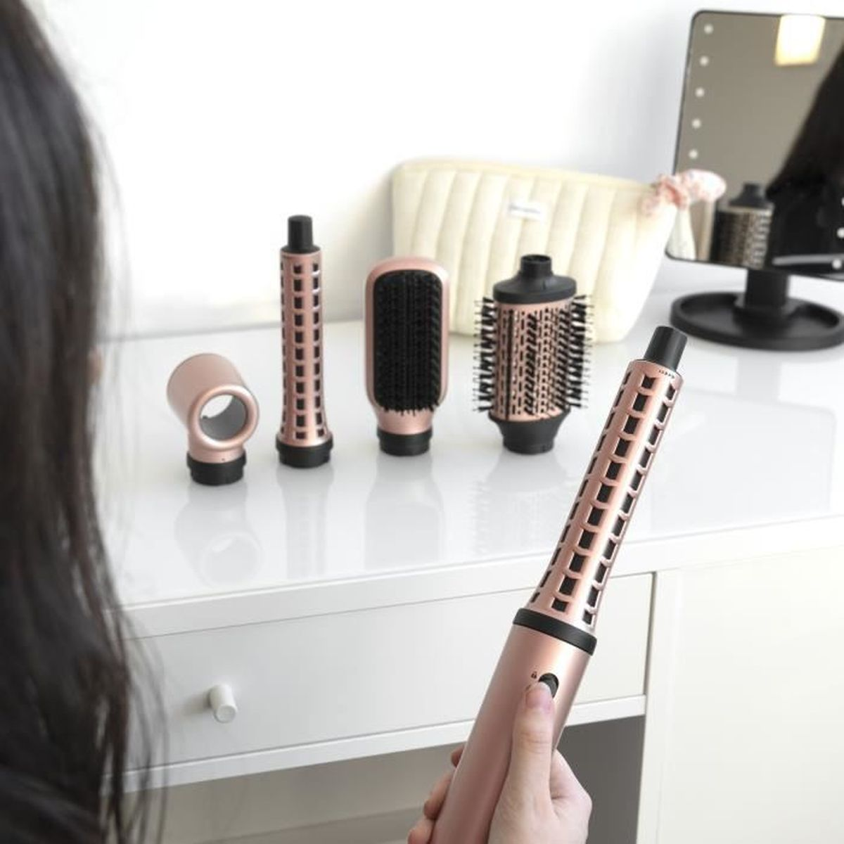 Una donna tiene in mano una piastra per capelli in oro rosa, con altri accessori su un tavolo bianco.