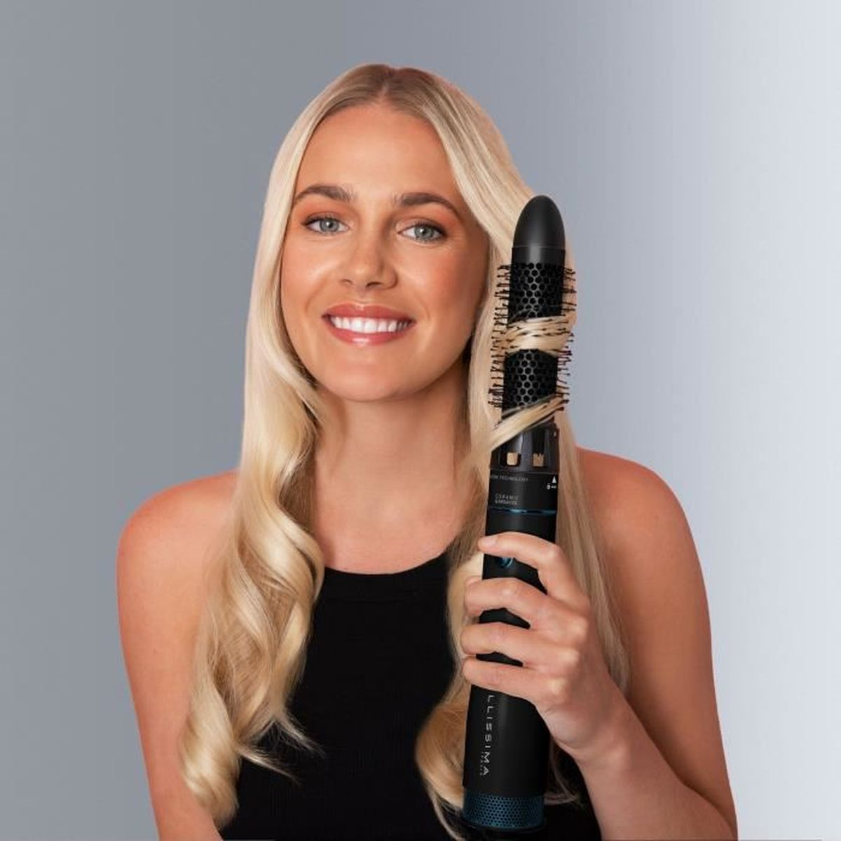 Una donna bionda sorride tenendo un styler nero con capelli arricciati.