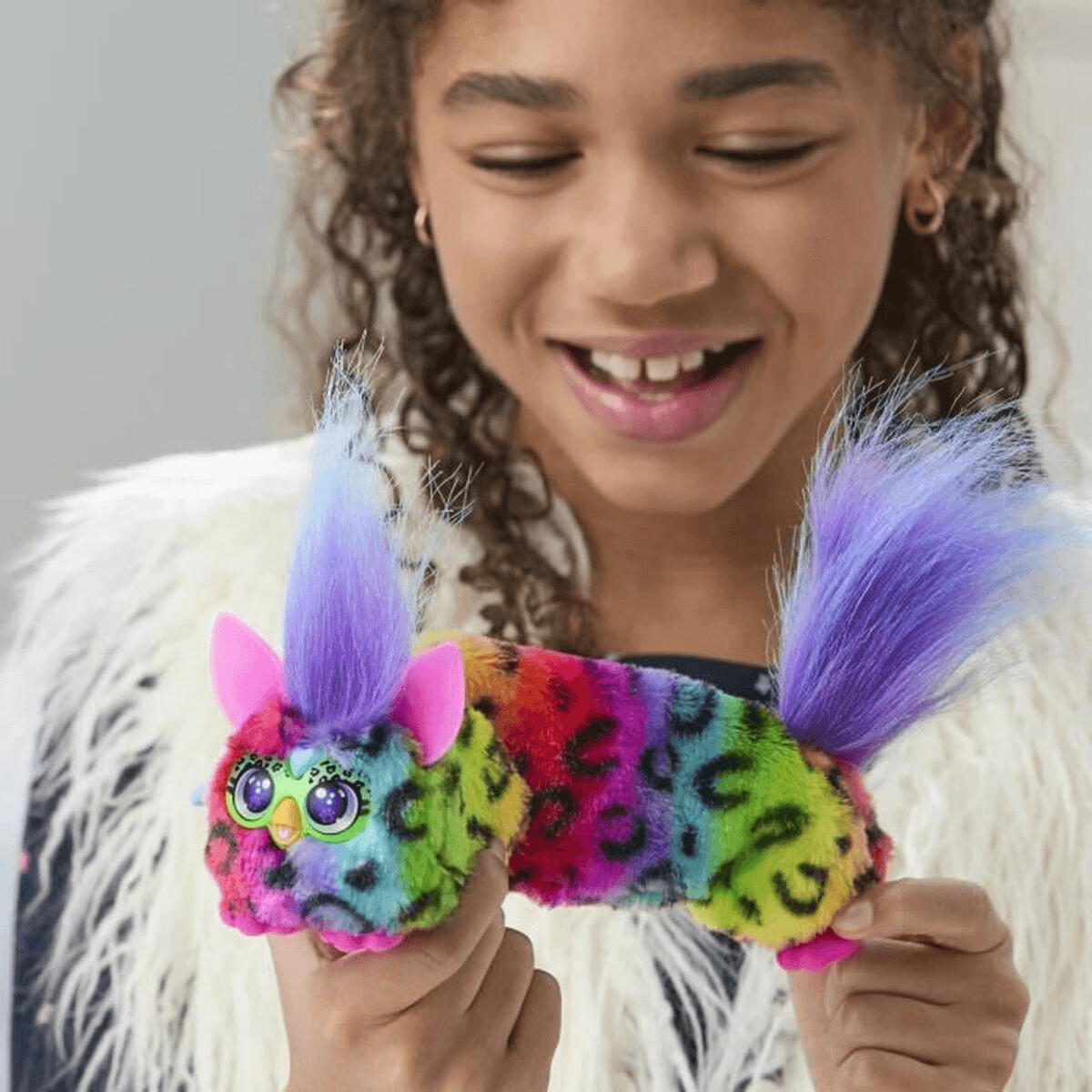 Una niña sonriente sostiene un peluche largo y colorido con pelo morado en la cabeza y la cola.