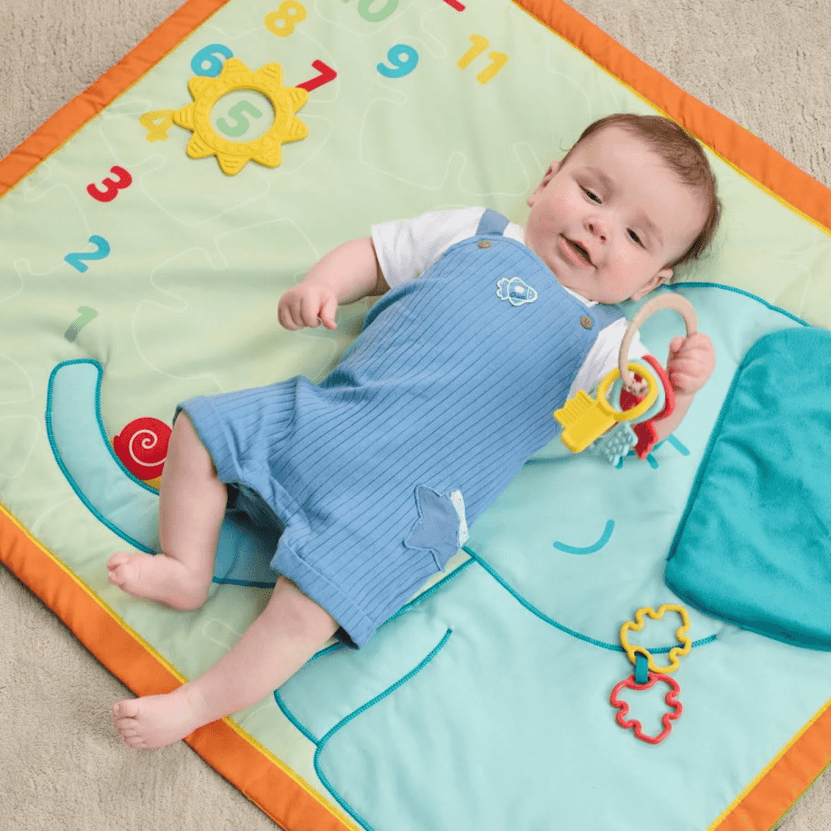 Un bebé feliz con peto azul yace en una alfombra de juegos colorida y sostiene un juguete.