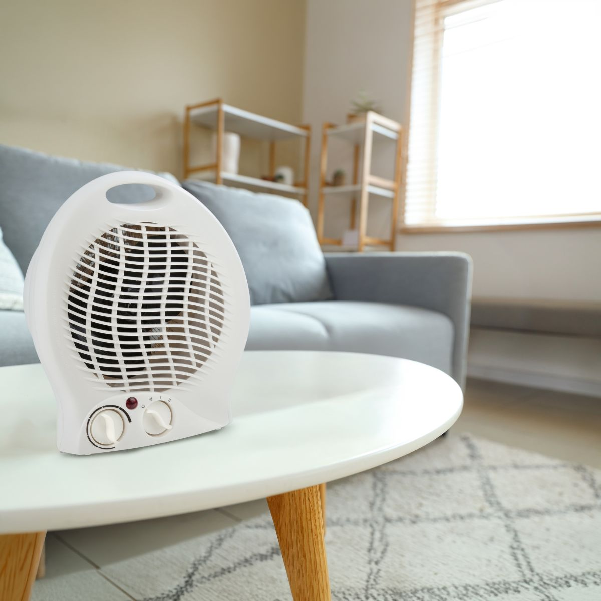 Un radiateur soufflant blanc est posé sur une table ronde dans un salon avec un canapé gris et des étagères.
