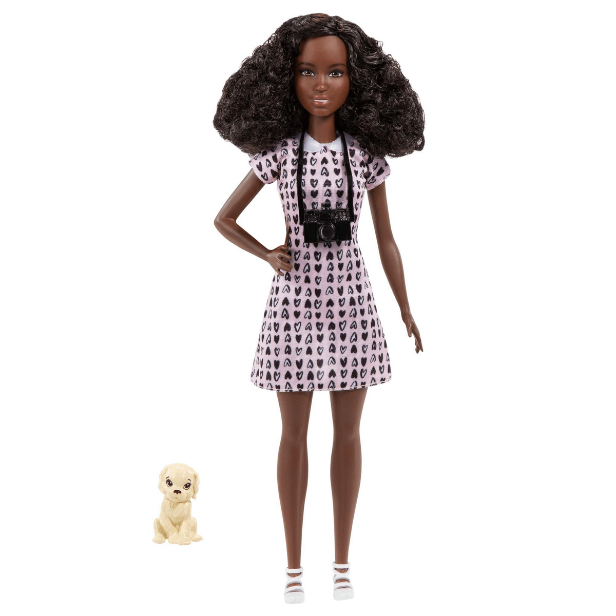 Una muñeca Barbie negra con cabello rizado que viste un vestido rosa con estampado de corazones, una cámara negra y un cachorro de juguete.