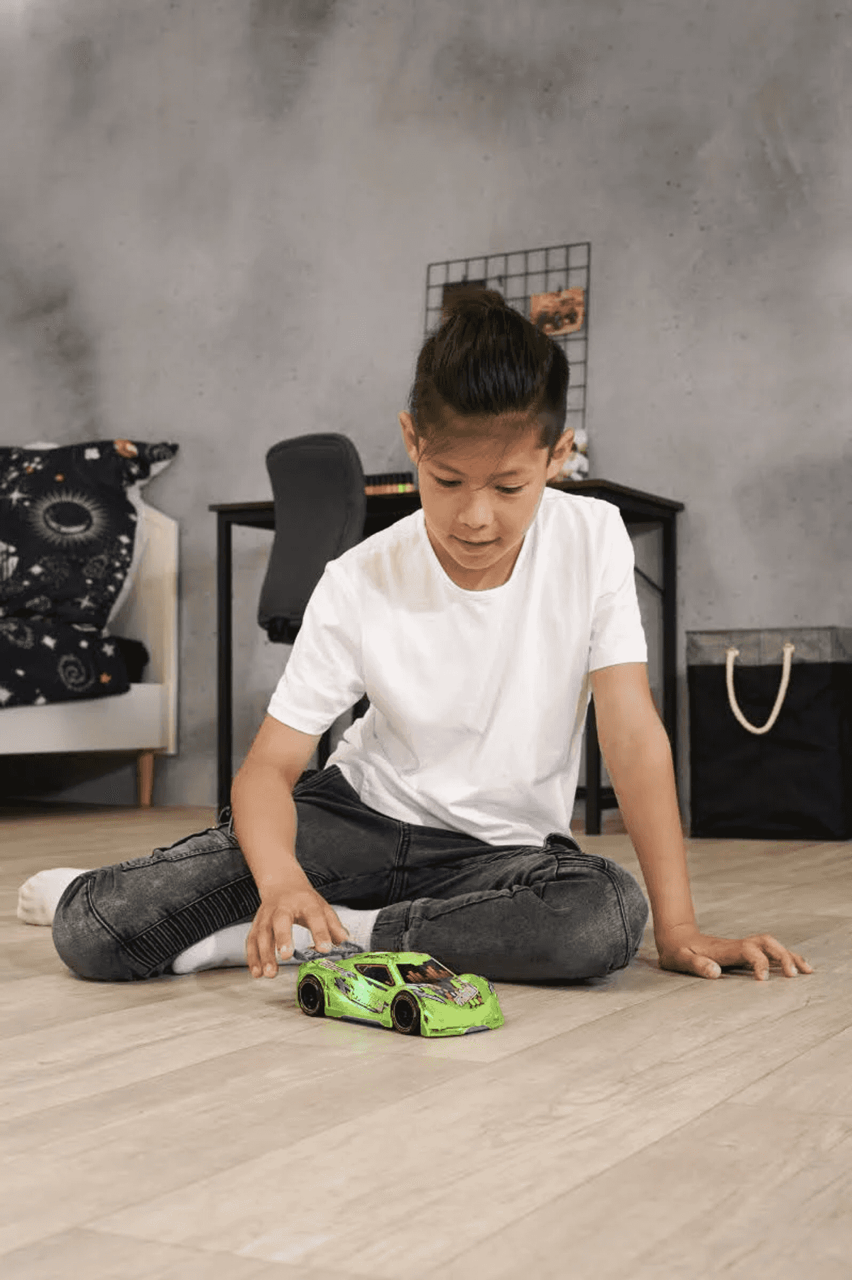 Un niño con una camiseta blanca y vaqueros oscuros juega con un coche de juguete verde en el suelo de madera.