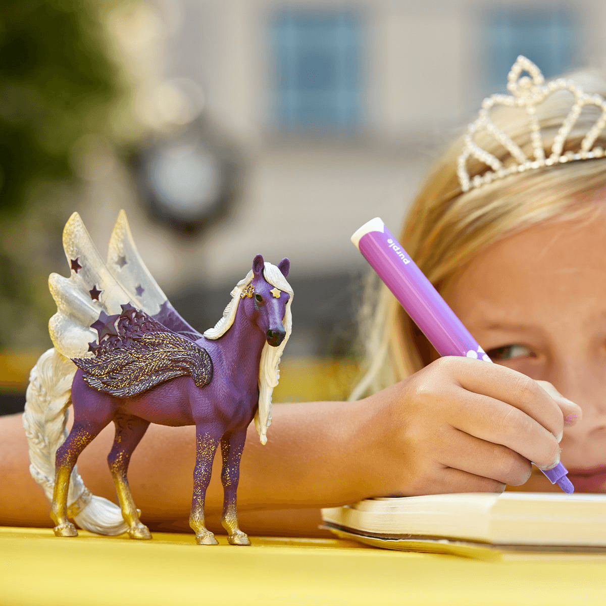 Ein blondes Mädchen mit Diadem malt neben einer lilafarbenen Pegasus-Figur.