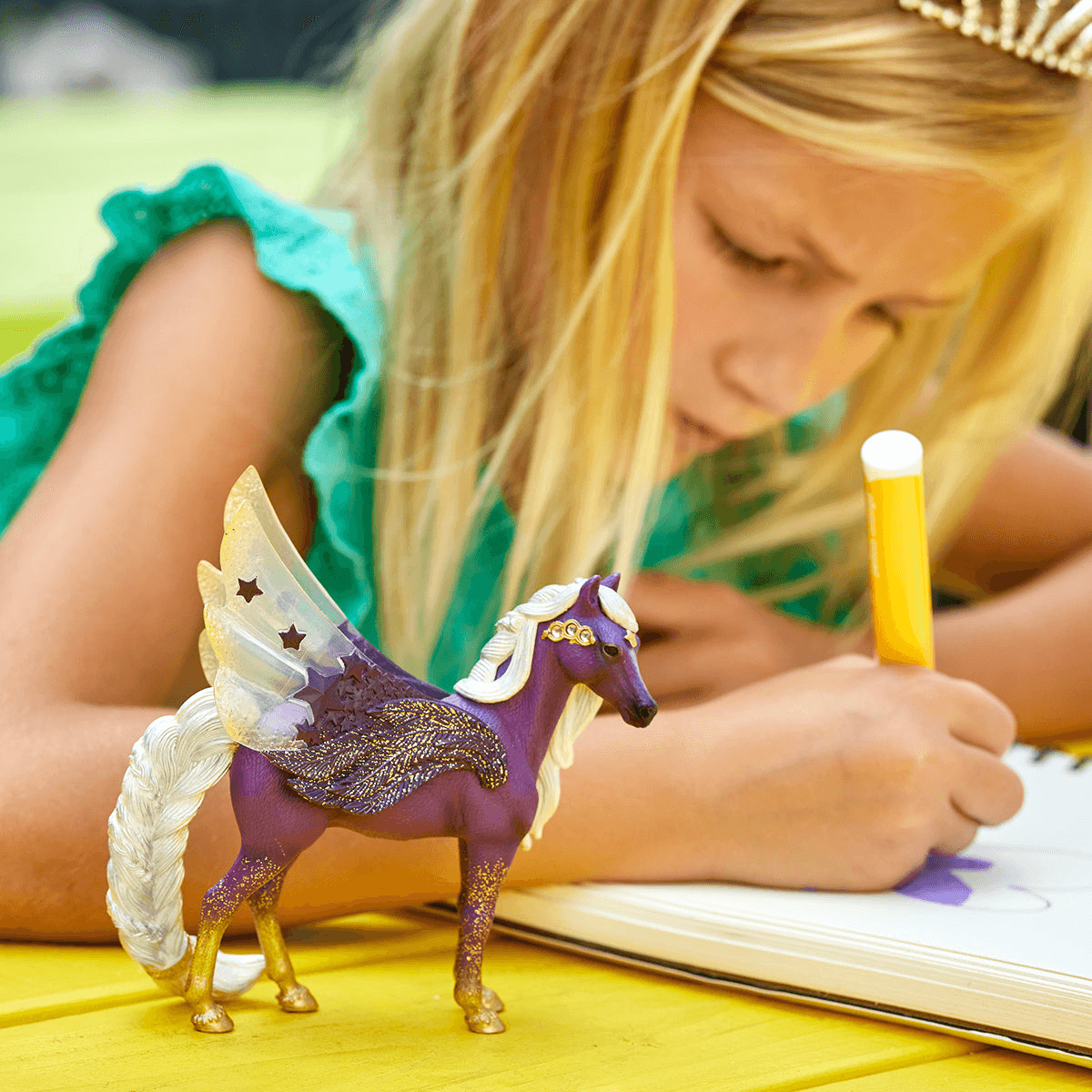 Ein blondes Mädchen in grünem Kleid zeichnet, während eine violette Pegasus-Figur mit goldenen Flügeln auf dem Tisch steht.