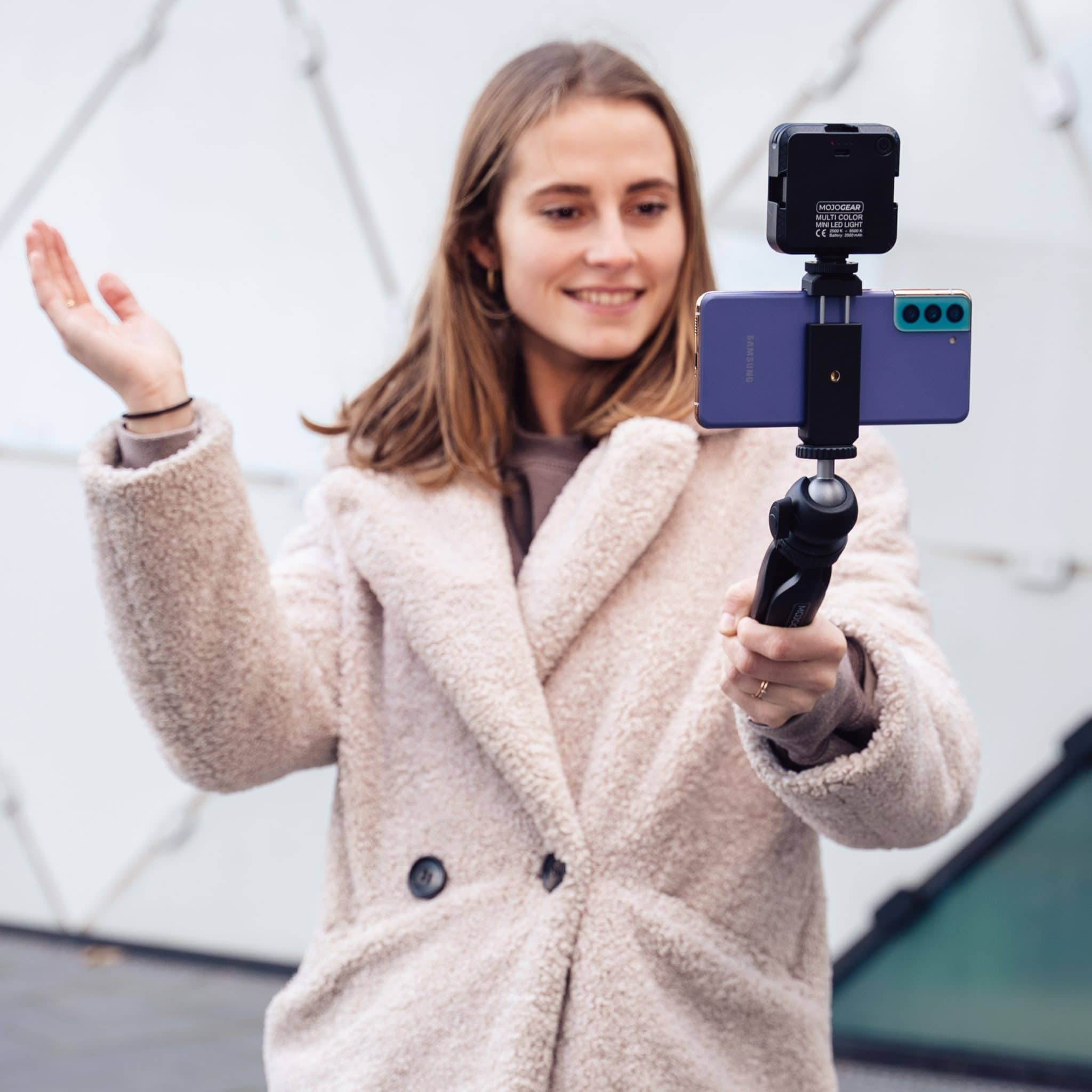Een vrouw in beige jas houdt een telefoontje op een statief, lacht en zwaait.