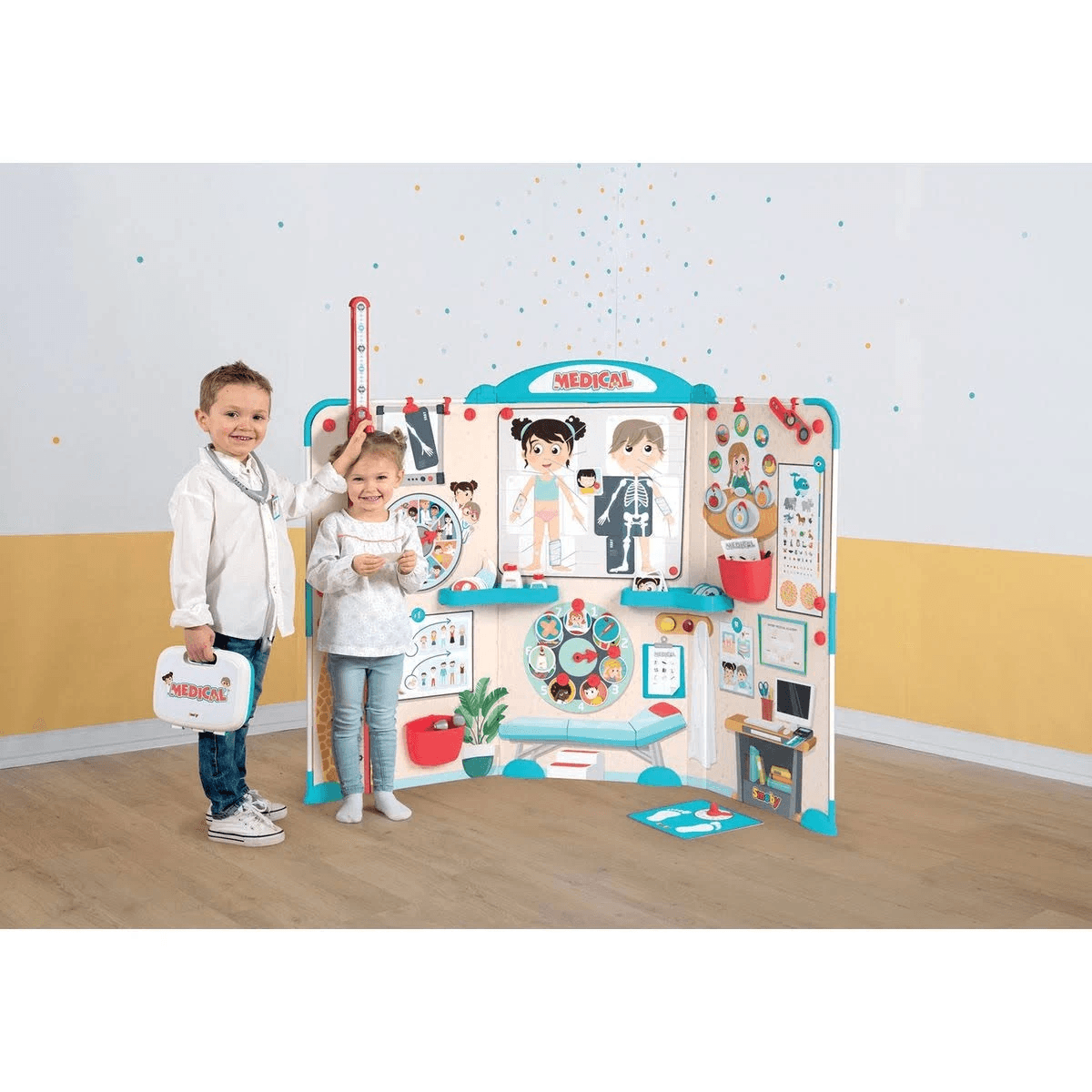 Dos niños juegan con un centro médico de juguete, con gráficos y equipos contra una pared.