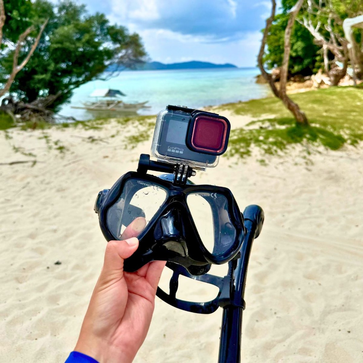 Een hand houdt een duikbril met GoPro-camera vast op een zandstrand met een tropische zee.