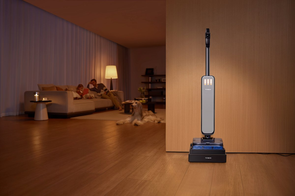 Een grijze steelstofzuiger staat op zijn oplaadstation op een houten vloer in een gezellige woonkamer.