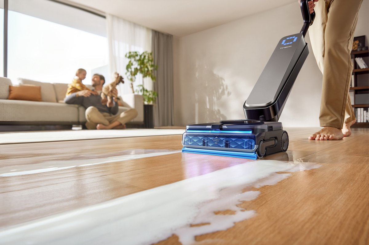 Une personne nettoie un liquide renversé avec un nettoyeur de sol bleu dans un salon, tandis qu'une famille est assise.