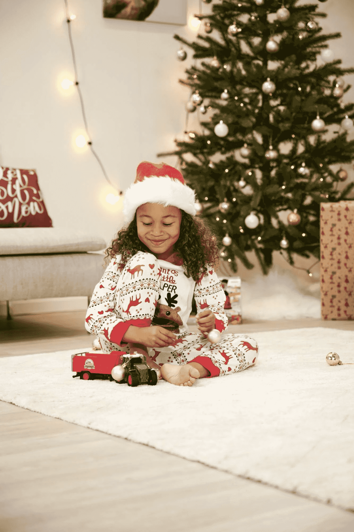 Una bambina in pigiama e cappello da Babbo Natale gioca con un camion giocattolo vicino a un albero di Natale.