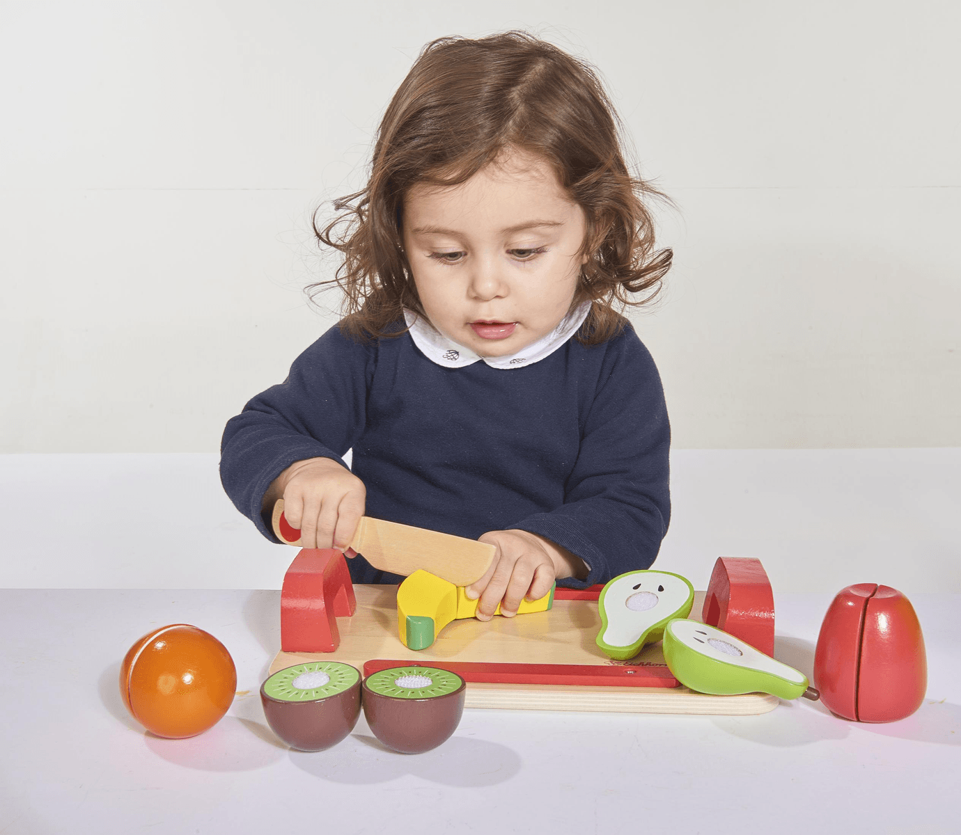 Un niño corta fruta de juguete de madera en una tabla, rodeado de naranjas, kiwis, aguacates y manzanas.