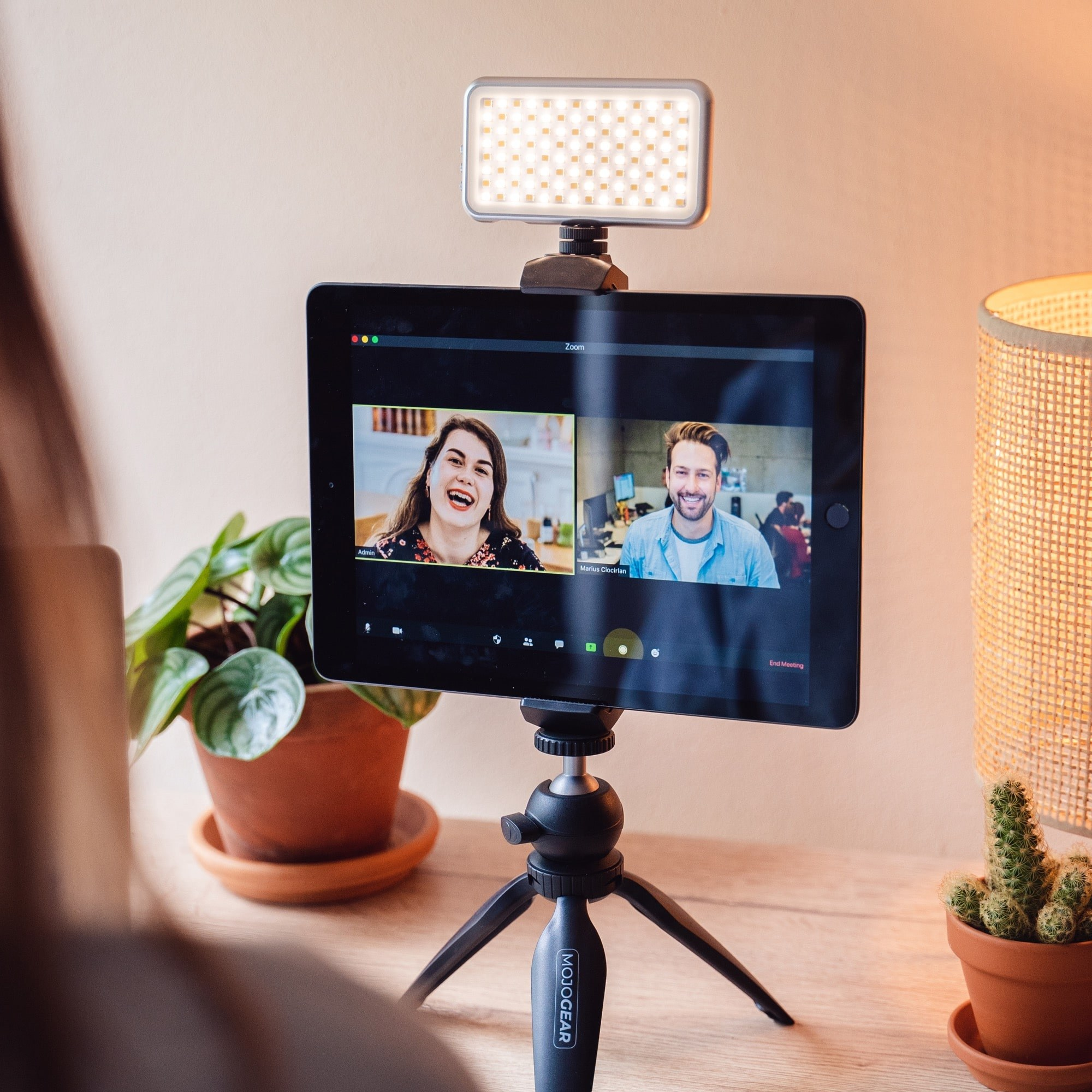 Une tablette sur trépied avec un appel vidéo de deux personnes, une lumière, une plante et une lampe sur un bureau.