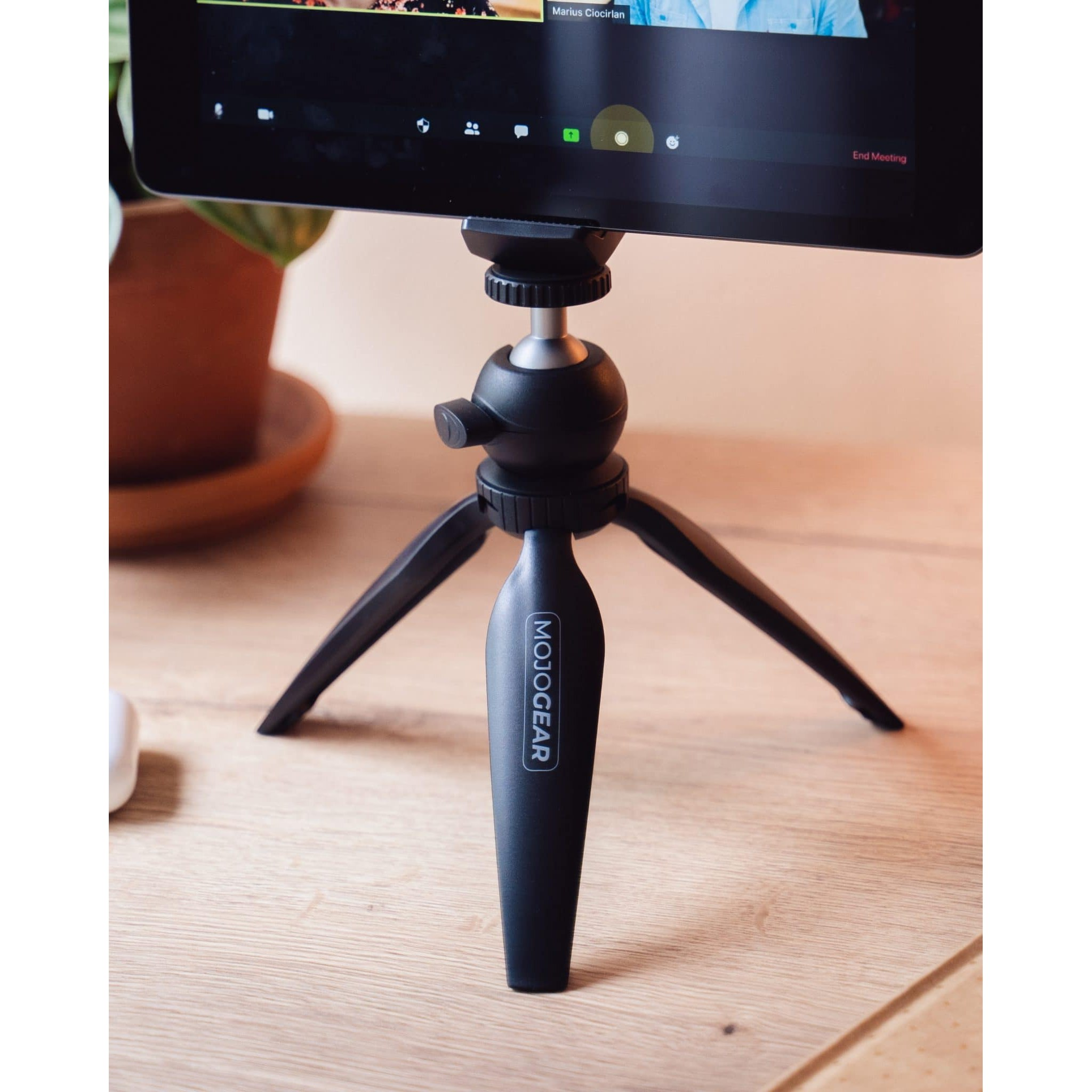 Un mini trépied noir Mojogear soutient une tablette sur un bureau en bois, avec une plante en pot en arrière-plan.