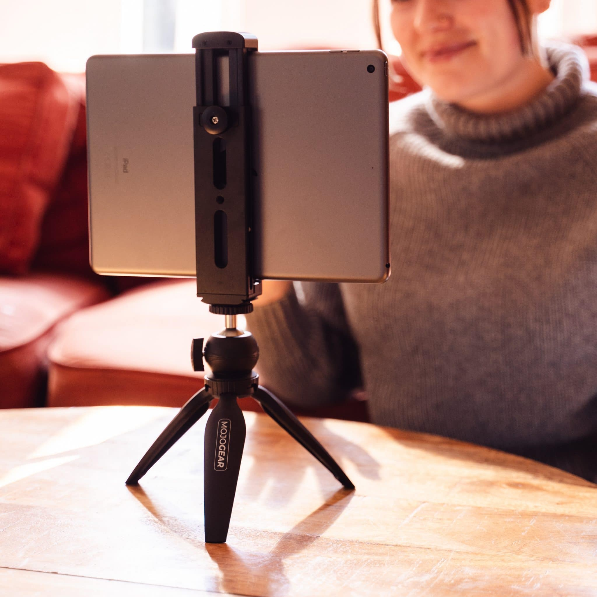 Une tablette dans un support sur un petit trépied en bois, avec une personne assise en arrière-plan.
