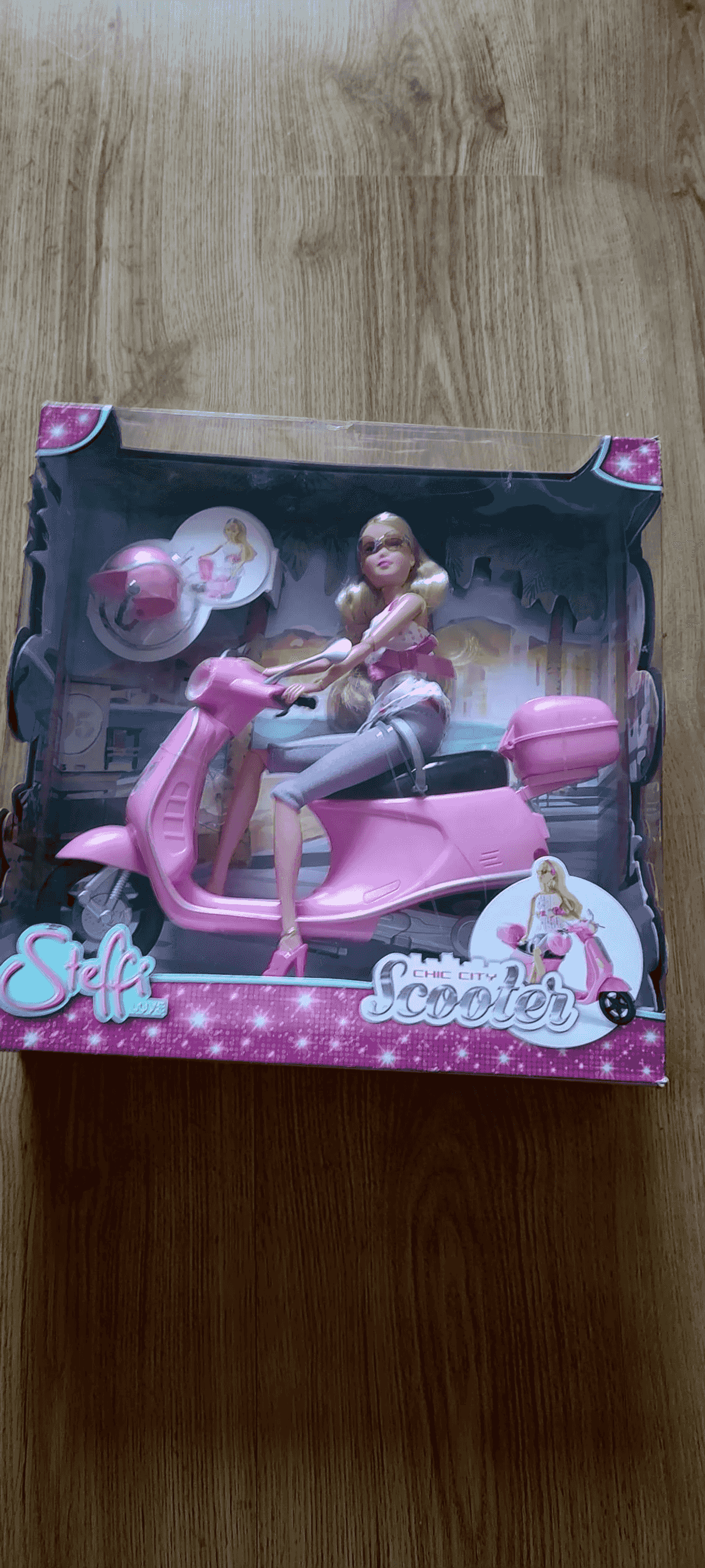 Eine blonde Modepuppe sitzt auf einem pinkfarbenen Spielzeugroller in einer bunten Verpackung.