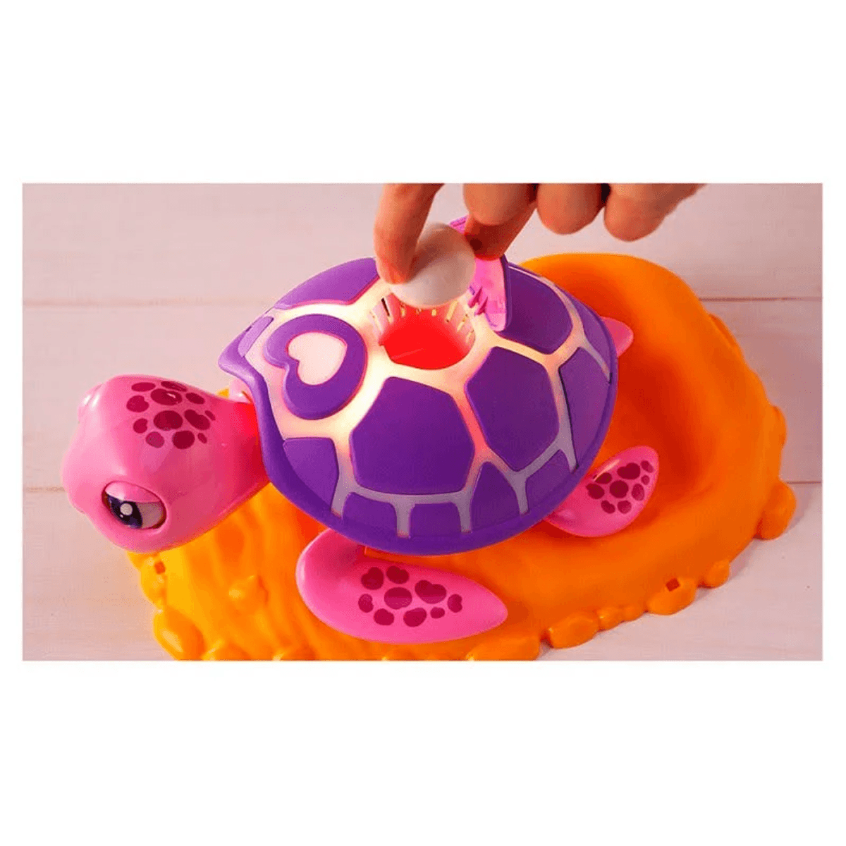 Una mano coloca un huevo blanco en una tortuga de juguete morada y rosa brillante sobre una base naranja.