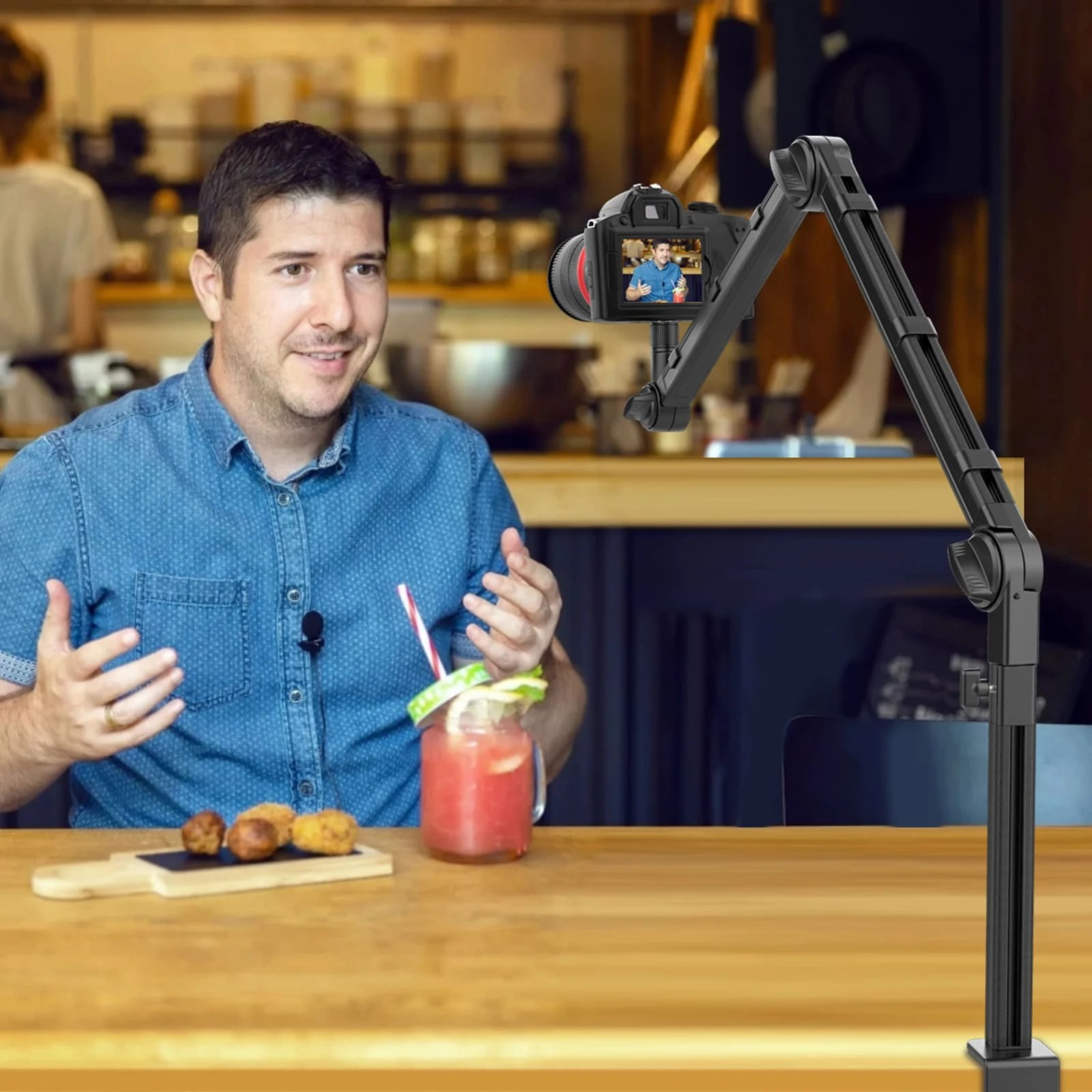 Un homme en chemise bleue est assis à une table en bois avec de la nourriture et une boisson, filmé par une caméra.