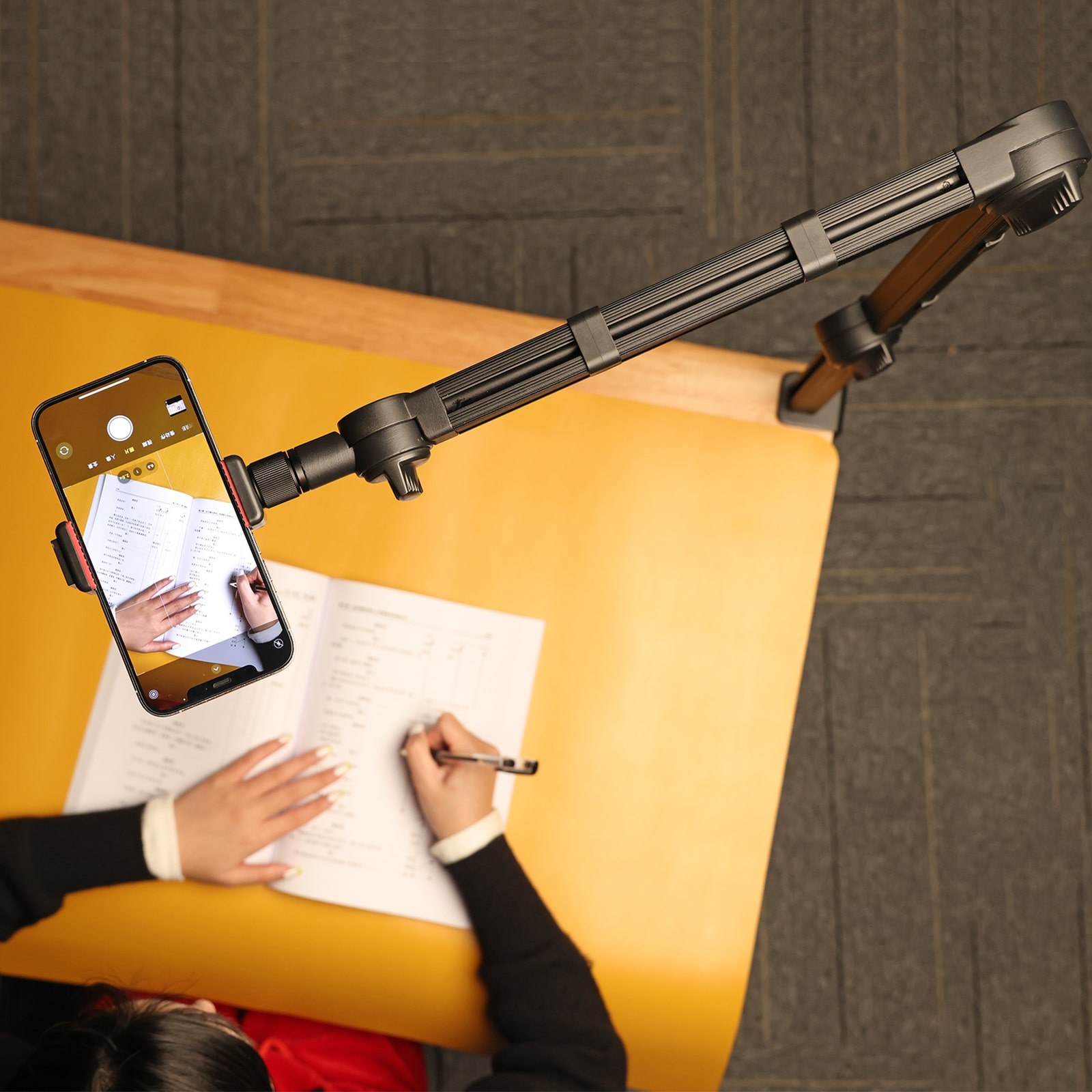 Vue en surplomb d'une personne écrivant dans un carnet sur un bureau jaune, filmée par un support de téléphone.