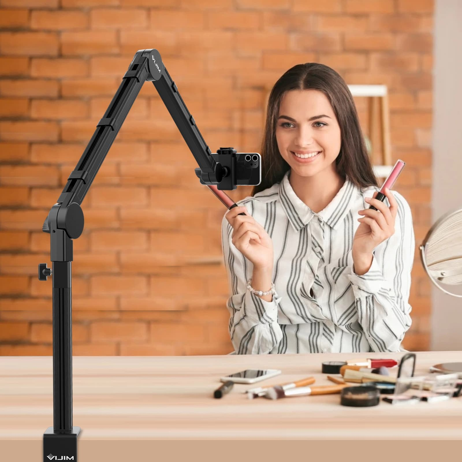 Une femme souriante tient du maquillage tout en filmant avec un smartphone sur un bras articulé.