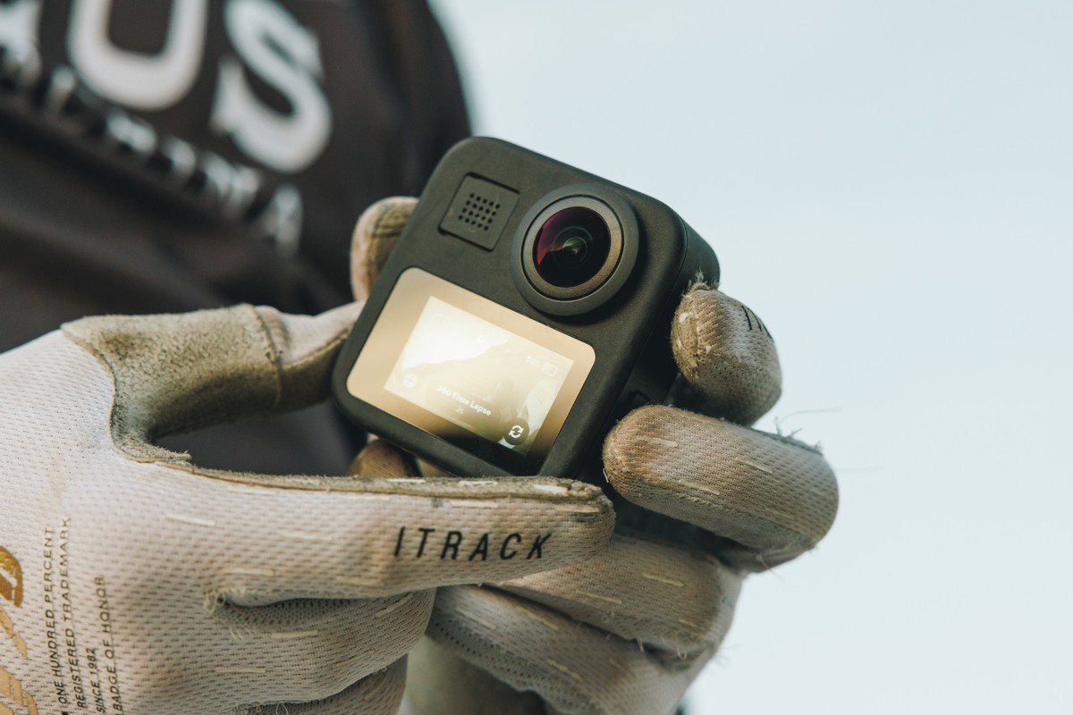 Mani guantate tengono una piccola action camera nera con schermo illuminato su sfondo sfocato.