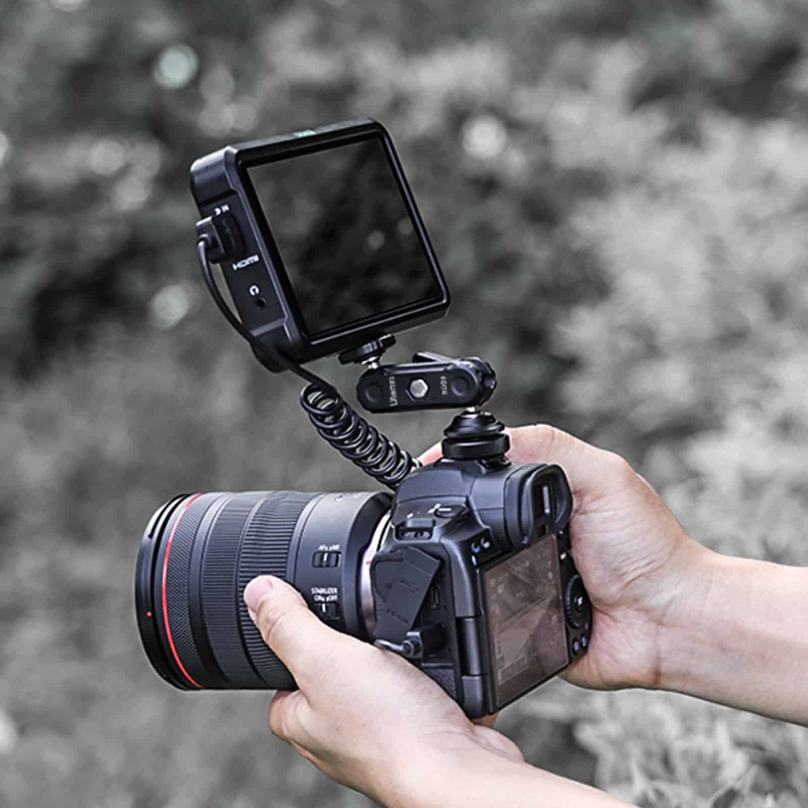Aparat DSLR z podłączonym zewnętrznym monitorem, trzymany w dłoniach na rozmytym tle.