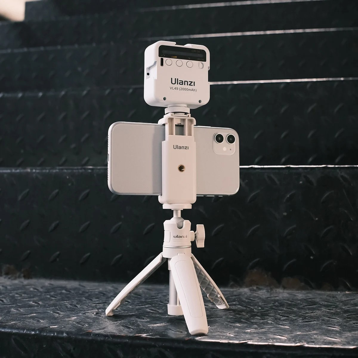 Un smartphone blanc sur trépied avec une lumière Ulanzi, devant des marches d'escalier en métal sombre.