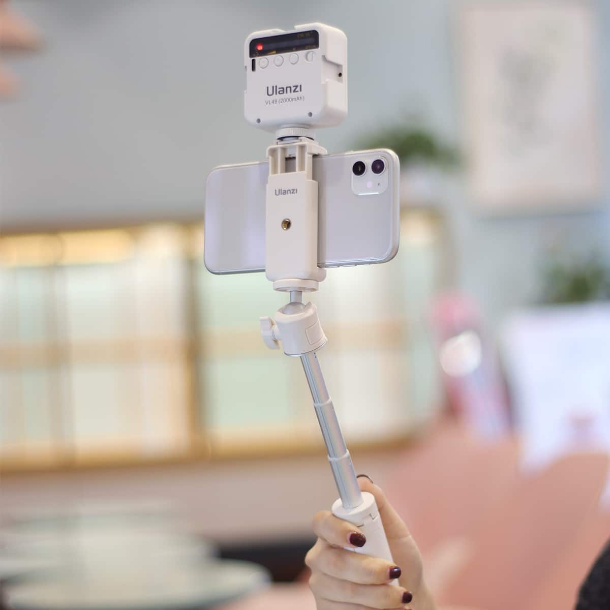 Une perche à selfie blanche tient un smartphone et une lumière LED Ulanzi, tenus par une main devant un fond flouté.