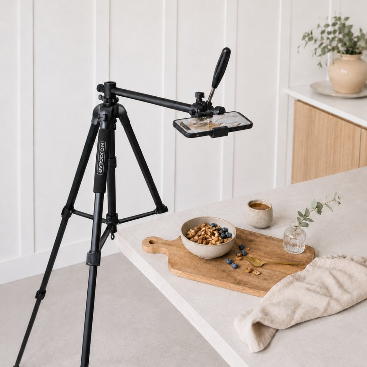 Un trépied noir avec un smartphone filme un bol de céréales et de myrtilles sur une planche en bois dans une cuisine lumineuse.