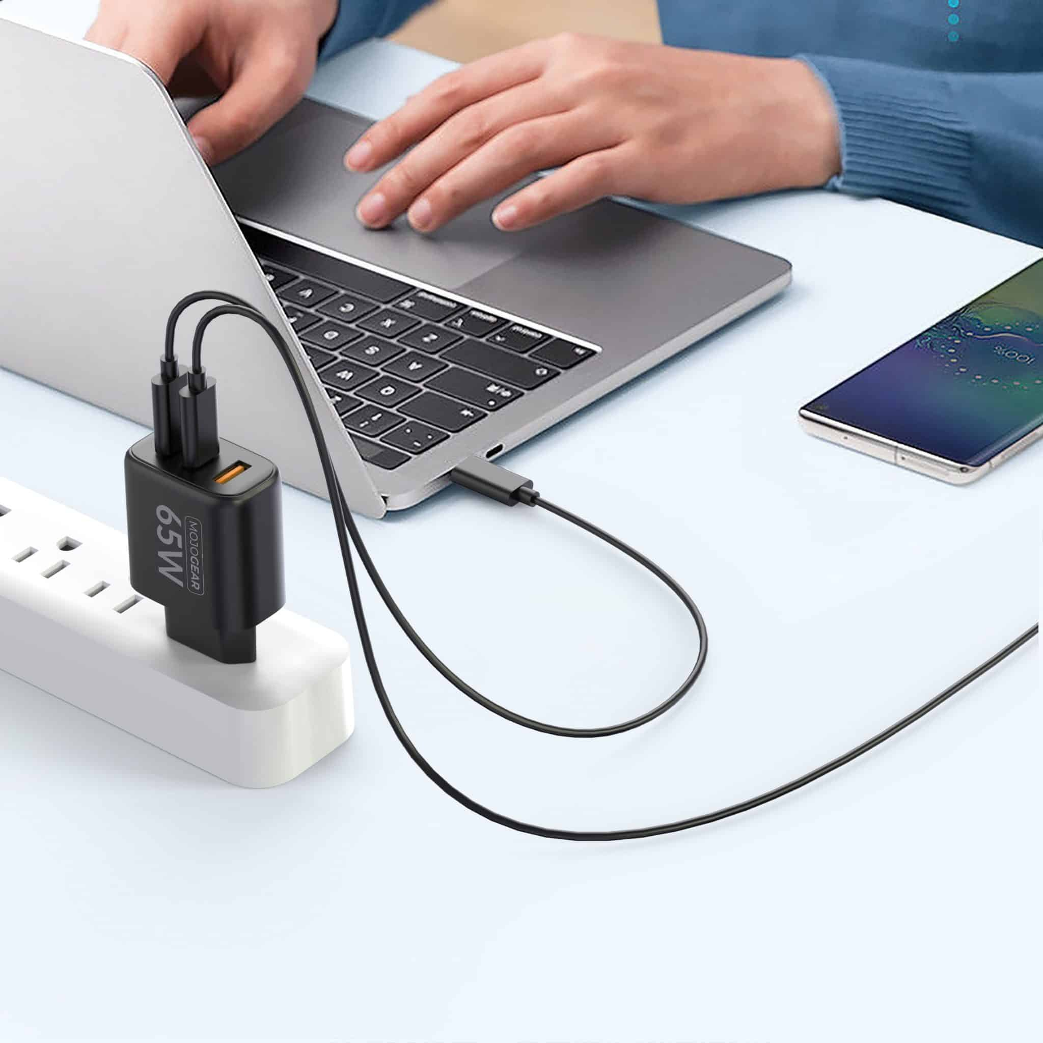 Un chargeur mural 65W branché avec des câbles noirs reliés à un ordinateur portable et un smartphone sur un bureau.