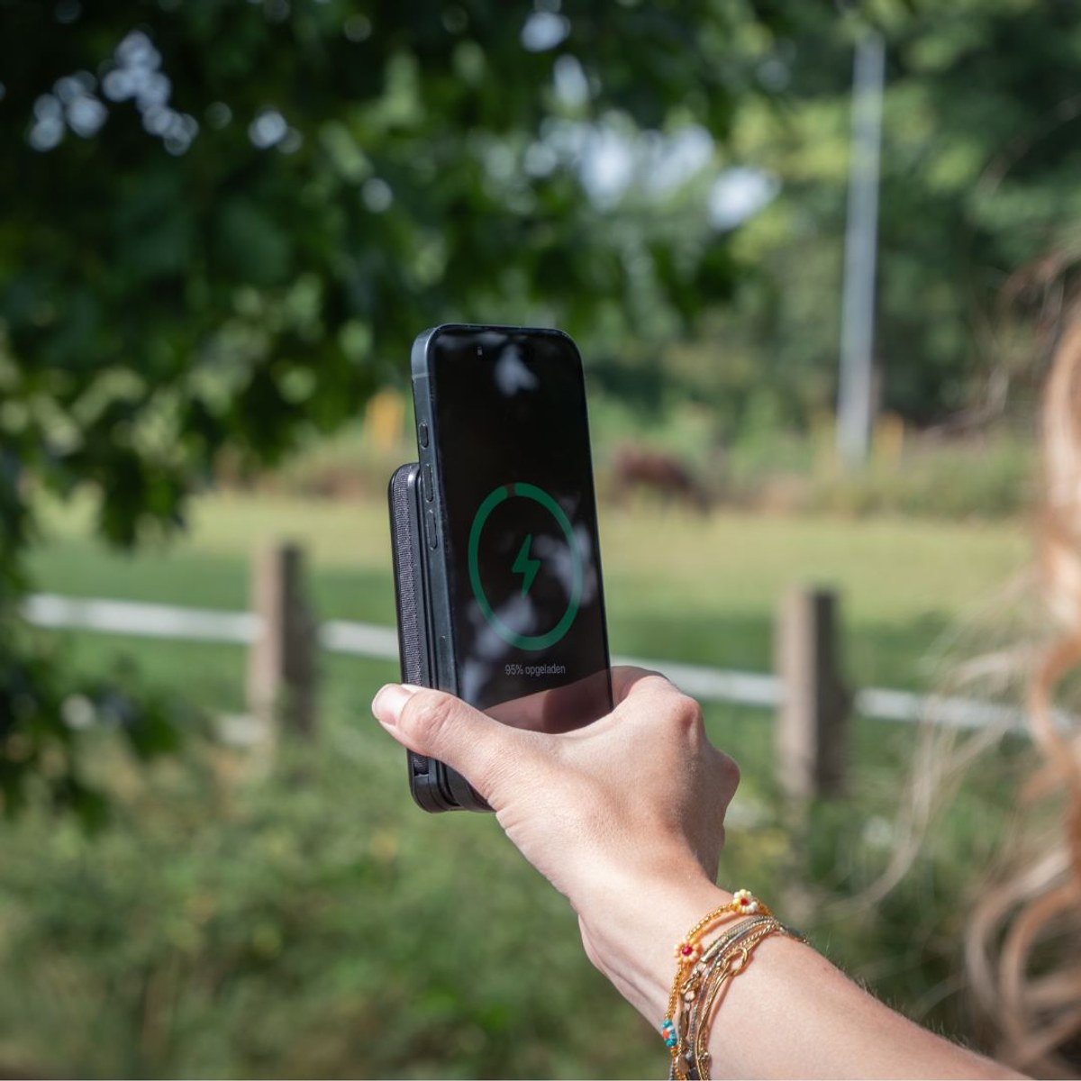 Eine Person hält ein Smartphone mit Ladesymbol im Freien, im Hintergrund grüne Wiese und Bäume.