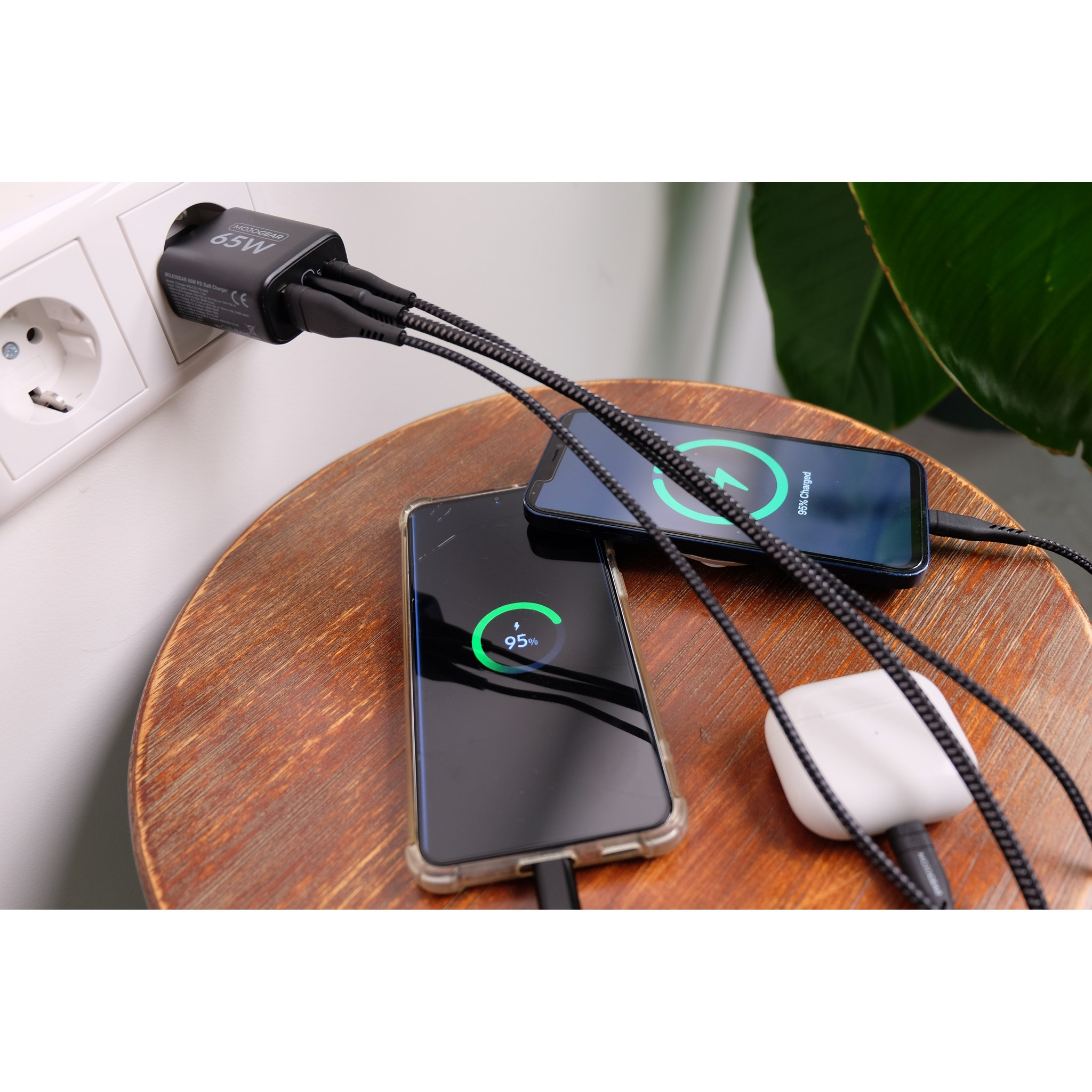 Ein 65W-Ladegerät lädt zwei Smartphones und Kopfhörer über schwarze Kabel auf einem Holztisch.
