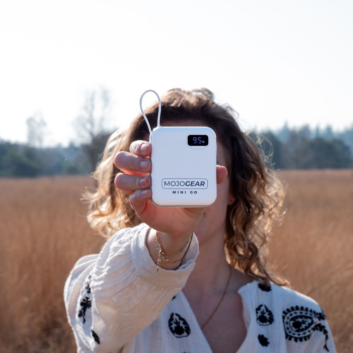 Eine Frau hält eine weiße Mojogear Mini Go Powerbank in die Kamera, im Hintergrund eine Wiese mit Bäumen.