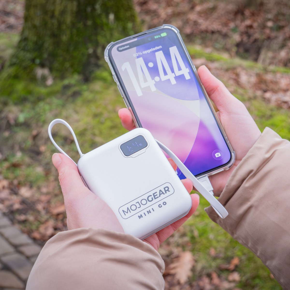 Eine Person hält eine weiße MojoGear Mini Go Powerbank im Freien, die ein Smartphone lädt.
