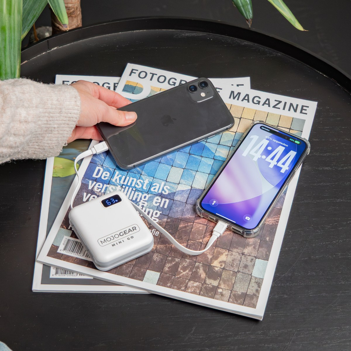 Eine Hand hält ein Handy, während eine weiße Powerbank ein anderes Handy lädt, das auf Zeitschriften auf einem Tisch liegt.