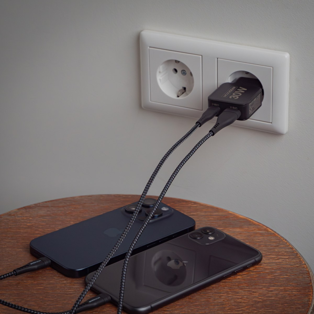 Two black iPhones on a wooden table, plugged into a wall USB charger.