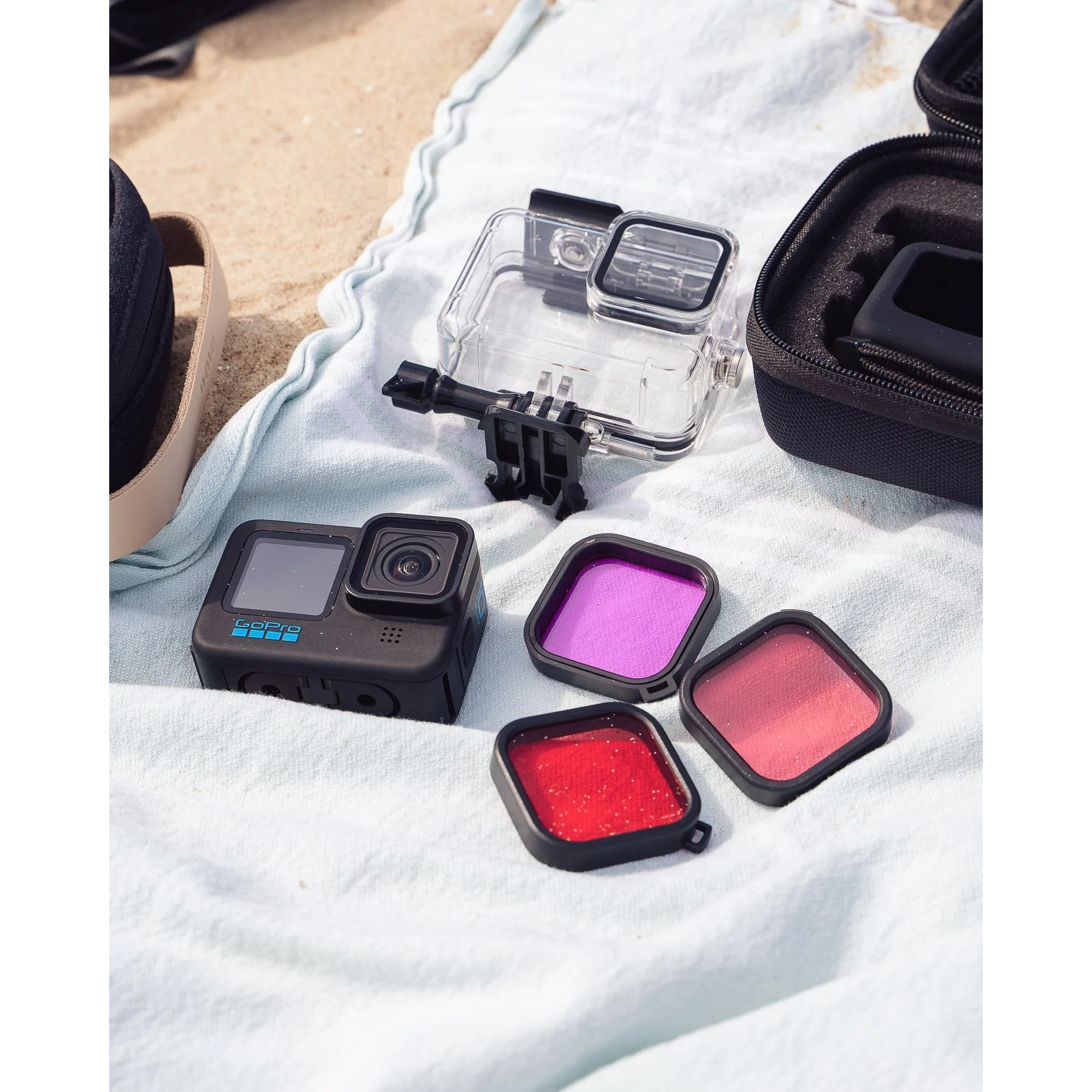 Een GoPro-camera, waterdichte behuizing, drie gekleurde lensfilters en etuis op een lichtblauwe handdoek.