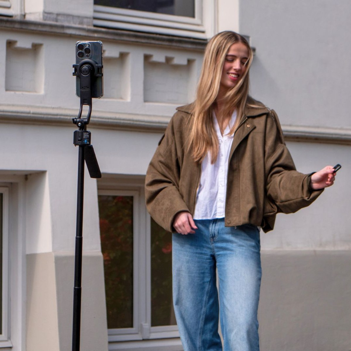 Een blonde vrouw in een bruine jas en jeans poseert buiten voor een smartphone op een statief.