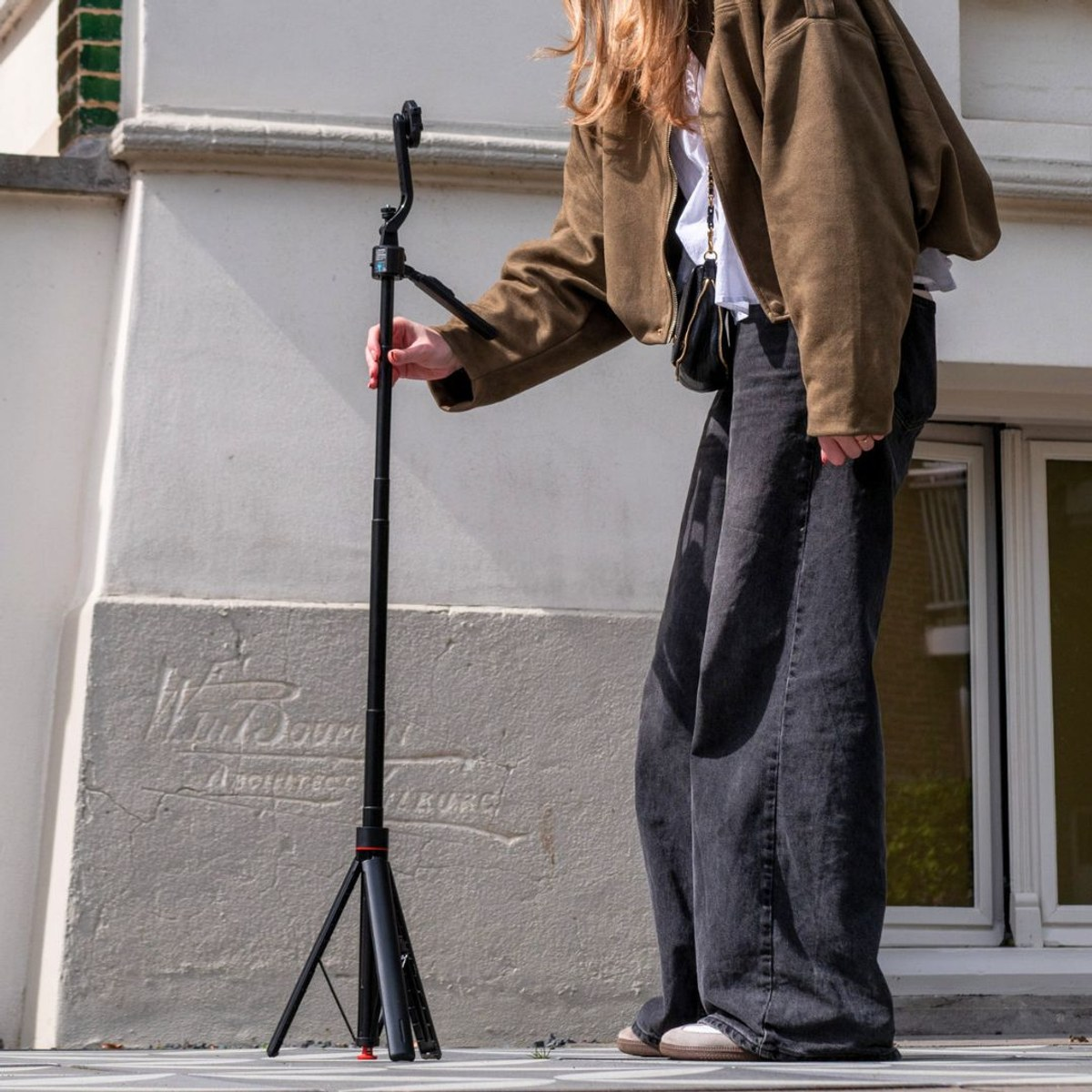 Iemand in een bruine jas en zwarte jeans stelt buiten een hoog zwart statief af.